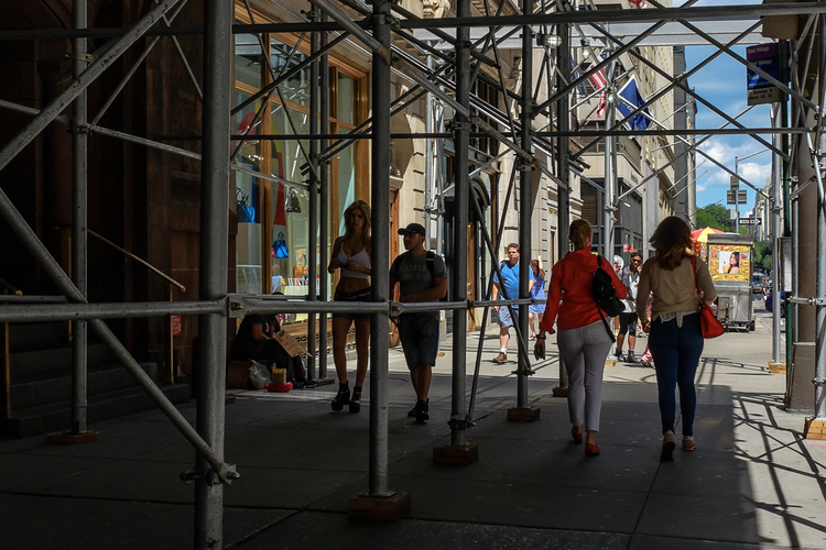 People stroll under the criss-crossed bars of scaffolding shading a New York City sidewalk.